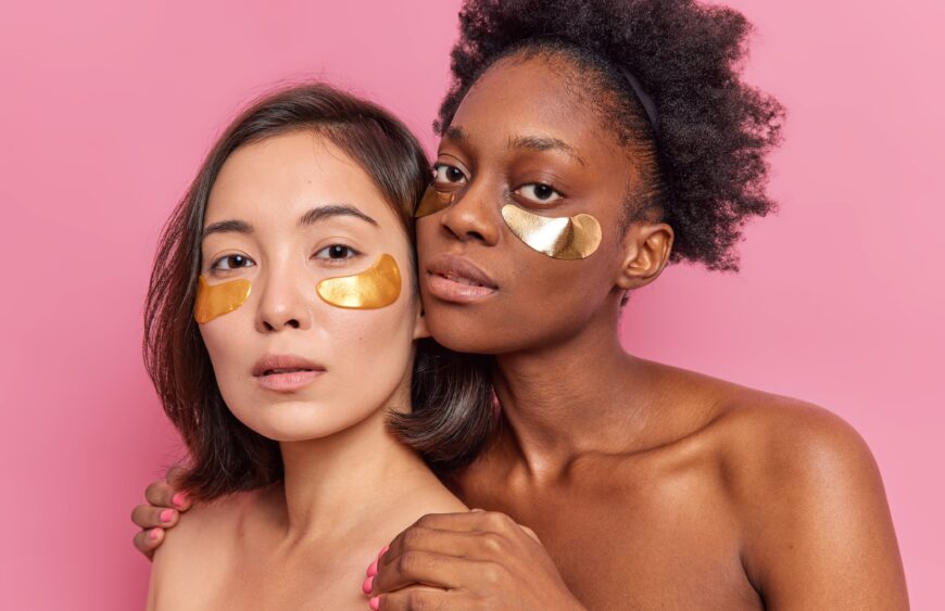 Two young multiethnic women apply golden patches under eyes stand closely to each other have healthy clean soft skin enjoy spa and beauty day isolated on pink wall. Face care treatment at home