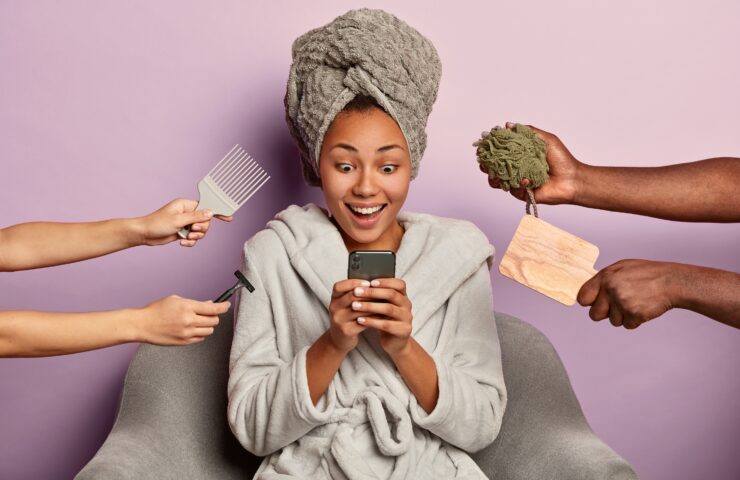 Joyful mixed race woman in domestic clothes gets message on cellphone, has shocked glad expression, four hands around with razor, mirror, comb and sponge, being groomed by professionals in salon